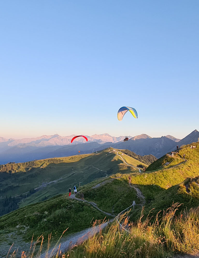 Zwei Paragleiter schweben bei Sonnenuntergang über grüne Alpen mit Wanderern und Bergstation im Vordergrund. | © Dorfgasteiner Bergbahnen AG