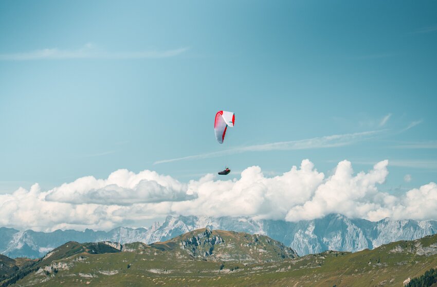 Ein einzelner Paragleiter fliegt bei Sonnenschein über grüne Berge mit Wolken und den Hohen Tauern im Hintergrund. | © Dorfgasteiner Bergbahnen AG