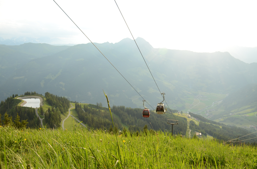 Zwei Gondeln schweben über grünen Almwiesen mit Speicherteich und weitem Bergpanorama im sonnigen Tal. | © Dorfgasteiner Bergbahnen AG