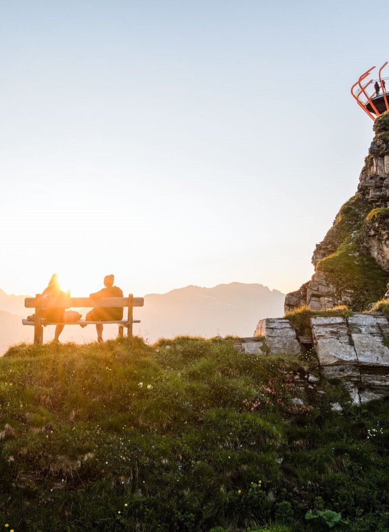 Paar auf Bank im Sonnenuntergang mit Blick auf die Berge, rechts die Plattform Glocknerblick auf grünem Felsen | © Gasteiner Bergbahnen AG