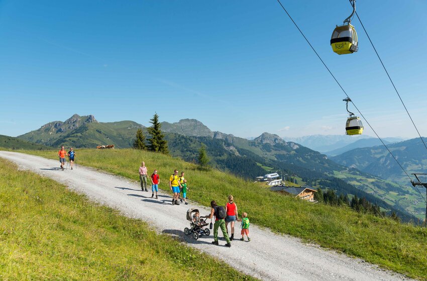 Familienwanderung im Großarltal mit Gondelblick auf das Tal und die umliegenden Berge | © Grossarler Bergbahnen GmbH & Co KG Wanderer mit Kindern und Kinderwagen auf Almweg in Großarl, gelbe Gondeln der Panoramabahn im Hintergrund | © Grossarler Bergbahnen GmbH & Co KG