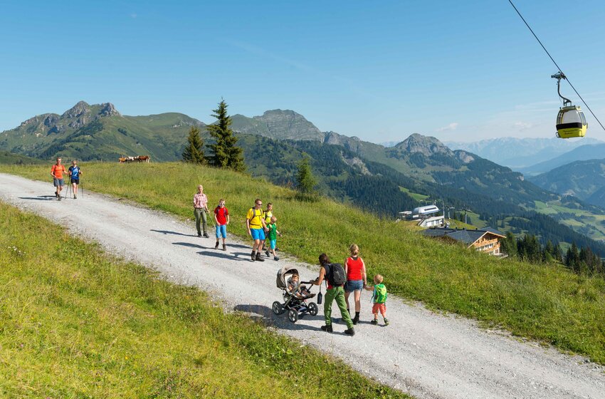 Familien mit Kindern und Kinderwagen auf sonnigem Almweg in Großarl, daneben gelbe Gondeln und Weitblick ins Tal | © Grossarler Bergbahnen GmbH & Co KG