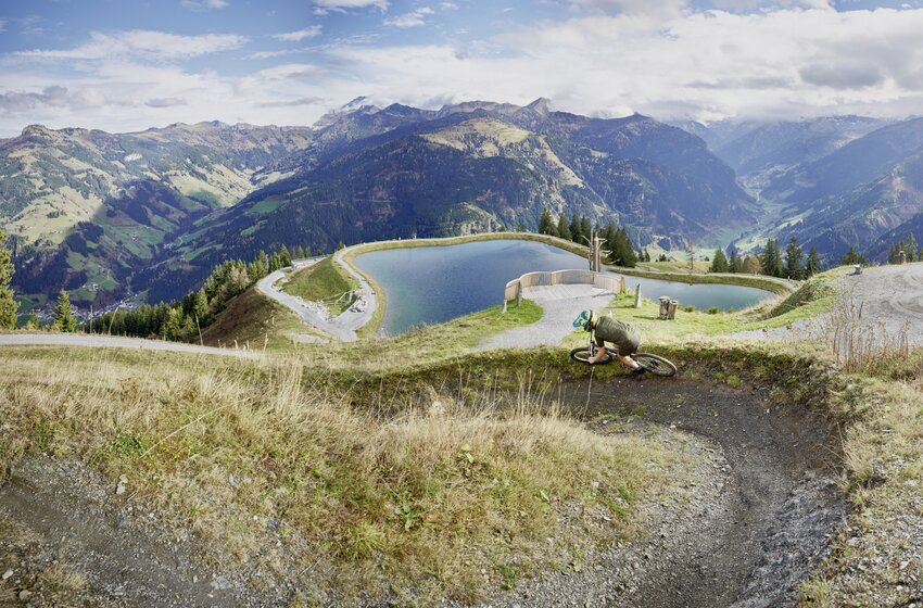 Mountain biker rides through a bend above a reservoir with a wide Alpine panorama beneath a partly cloudy sky. | © Großarler Bergbahnen GmbH & Co KG