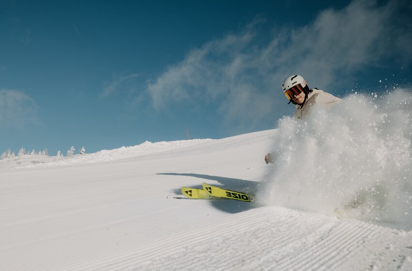 A skier carves a turn on a freshly groomed piste, spraying snow beneath a clear blue winter sky. | © Elisabeth Toferer