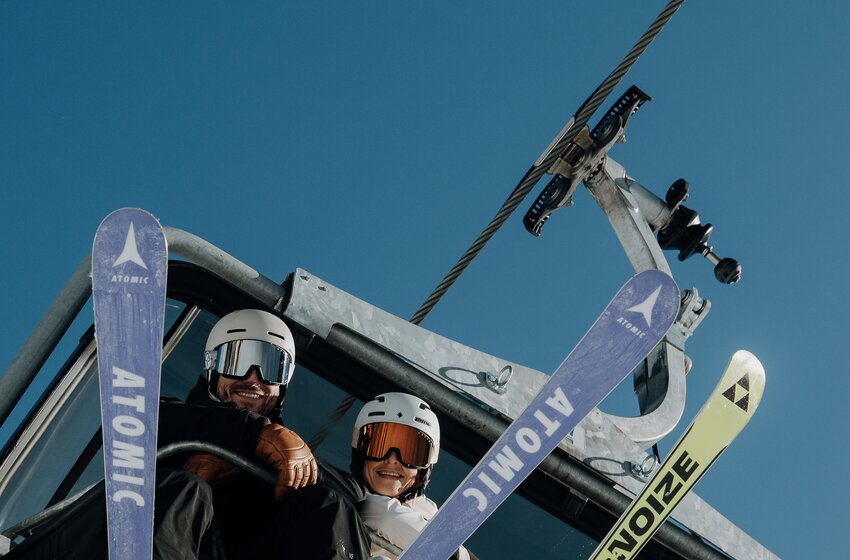 Two skiers sit on a chairlift with skis attached beneath a clear blue sky, looking down from above. | © Elisabeth Toferer