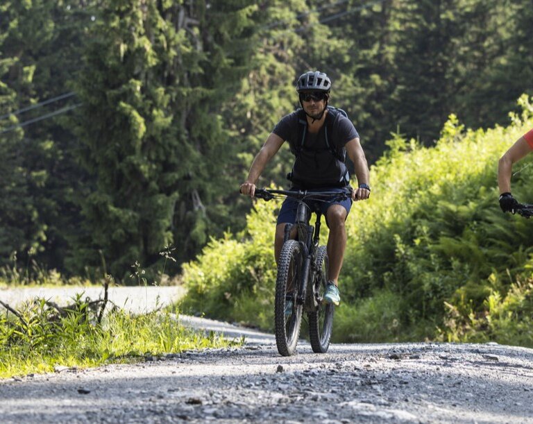 Two mountain bikers ride a sunny gravel trail through the lush forest landscape in Gastein valley. | © Gasteiner Bergbahnen AG