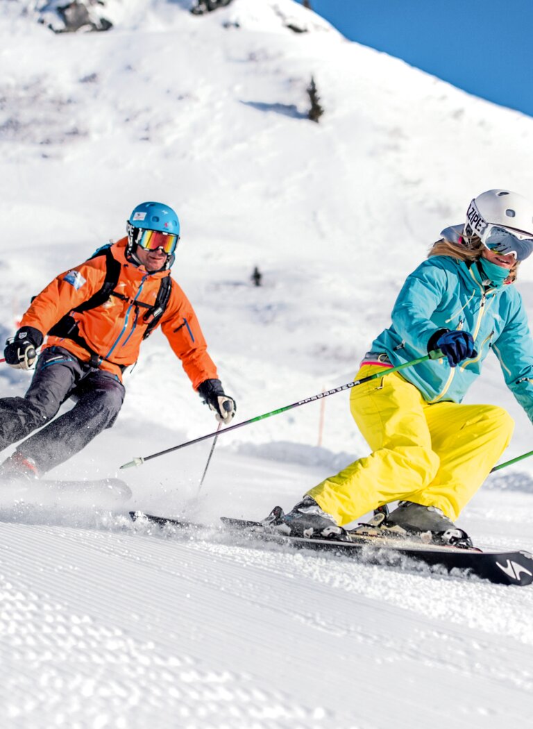 Two skiers carve down a wide, freshly groomed slope in sunshine and powder snow. | © Gasteiner Bergbahnen AG
