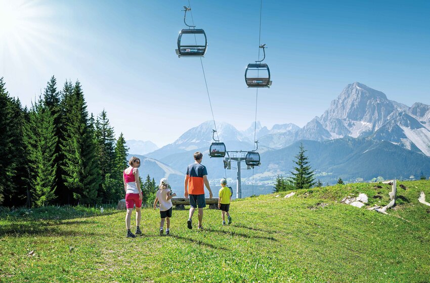 Family hiking through green alpine meadow with gondola and mountain backdrop on a sunny day. | © Ski amadé GmbH