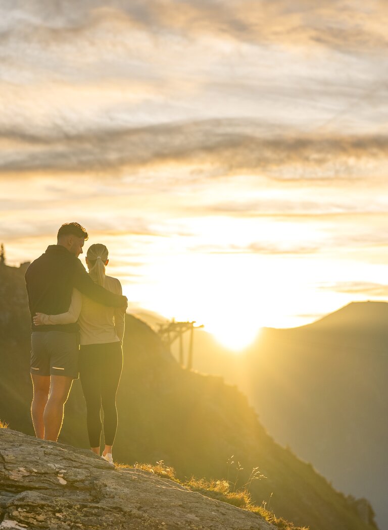 Couple stands closely embraced on a rock at the mountain summit, mountains and glowing sky in background | © Gasteiner Bergbahnen AG