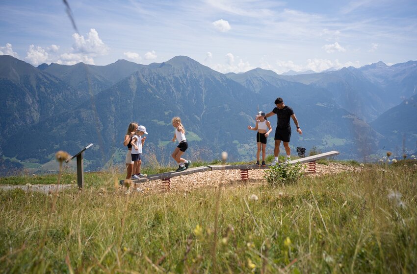 Familie mit Kindern balanciert auf Holzspielgerät auf Bergwiese, Alpenpanorama und Tal im Hintergrund | © Gasteiner Bergbahnen AG
