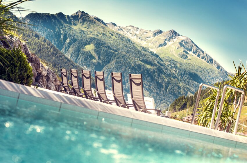 Several sun loungers line the pool edge overlooking green mountains and forested slopes beneath a clear sky | © Max Steinbauer A 5640 Bad Gastein