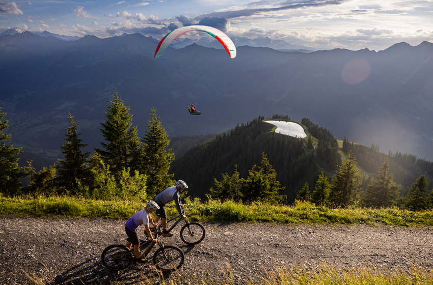 Zwei Mountainbiker fahren auf Schotterweg am Hang, darüber fliegt ein Paraglider, im Tal liegt ein Bergsee | © Gasteiner Bergbahnen AG