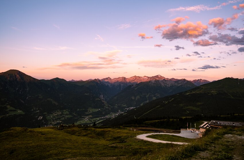 Mountain landscape at sunset with green valley, winding road in foreground and mountain range on horizon | © Gasteiner Bergbahnen AG